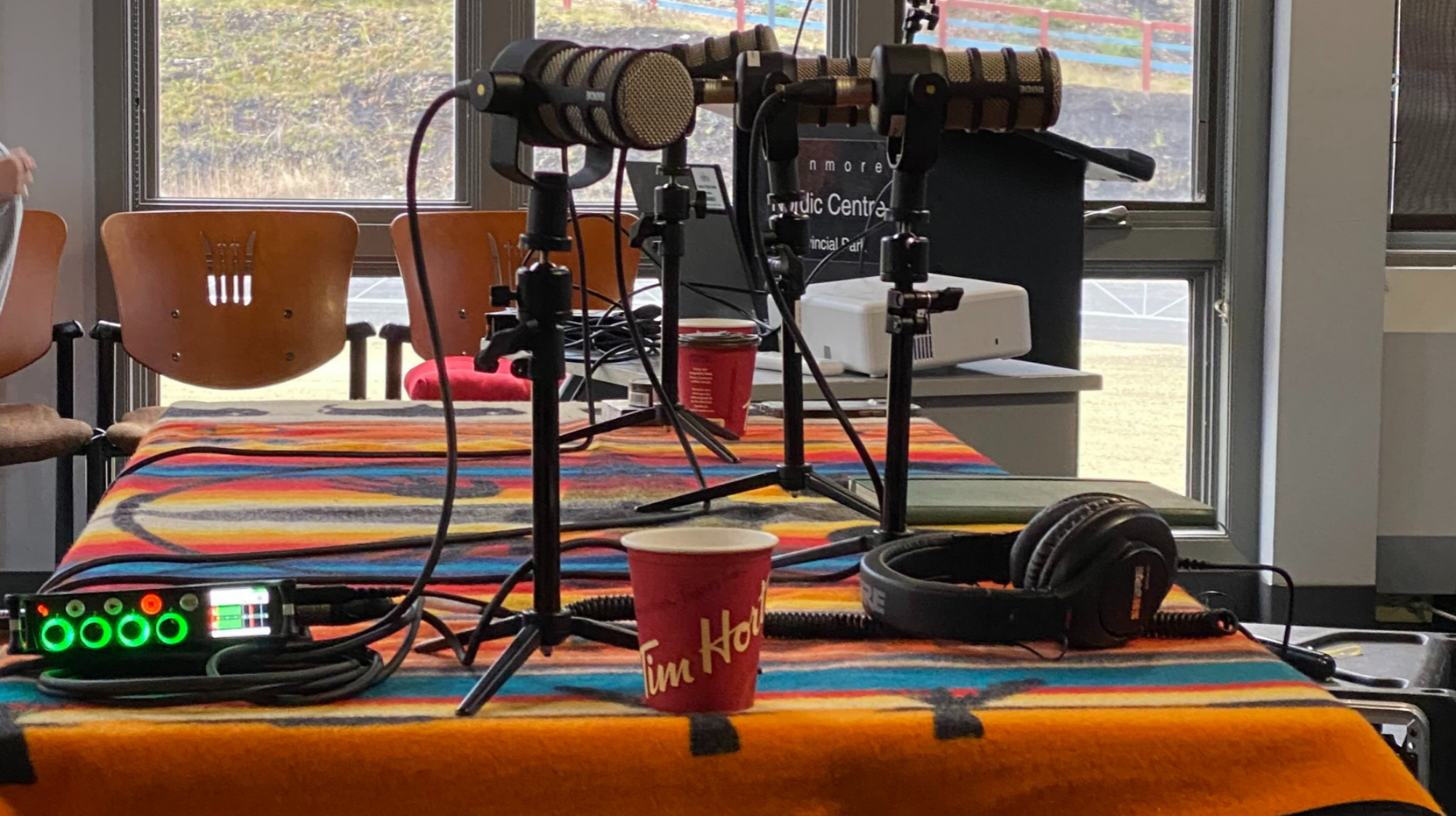 Image of podcast recorder and microphones on a table with a brightly-coloured blanket with Indigenous patterns. An empty Tim Horton's take out cup sits on the table among the gear. The windows in the back show a grassy landscape.